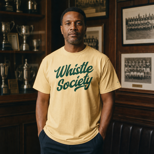 Man wearing the butter Ref' Crew Whistle Society Tee in a wood-panelled club room with trophies and vintage team photographs – forest-green vintage script across the chest