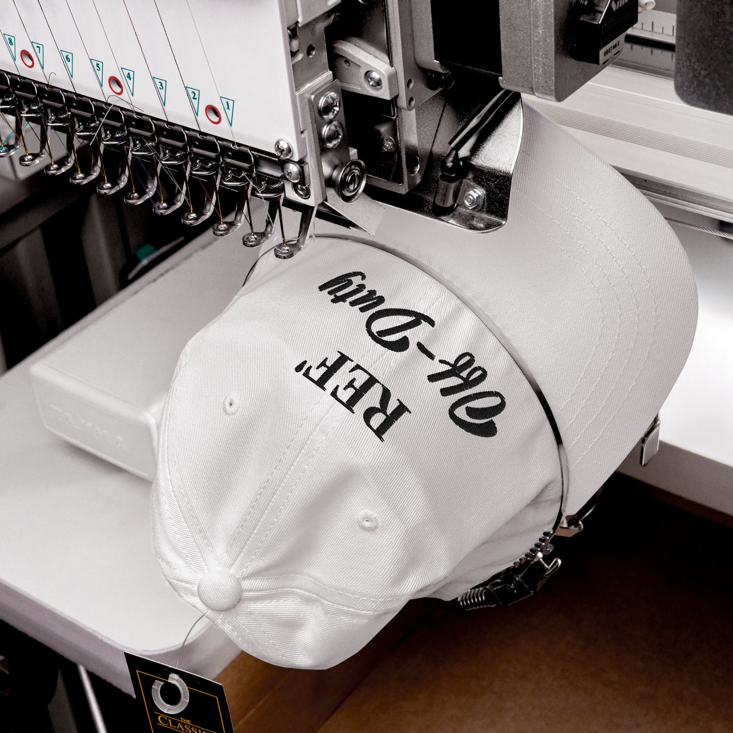 White "REF' Off-Duty" dad hat with black embroidery, being stitched on an industrial machine.