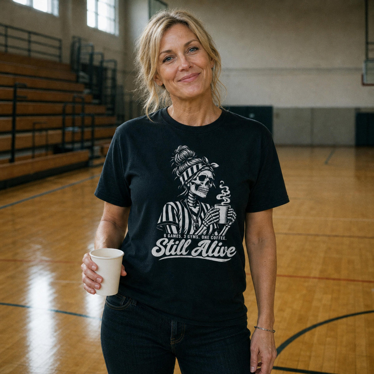 Woman holding a coffee cup in an empty gym, wearing the Black Still Alive Tee with the skeleton referee and "6 GAMES. 3 GYMS. ONE COFFEE. Still Alive" print – Ref' Crew basketball referee tee