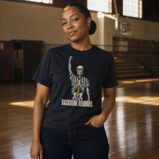 Woman wearing the Black Backbone Required Tee in an empty sunlit gym, skeleton referee print with fist raised and gold spine – Ref' Crew basketball referee tee