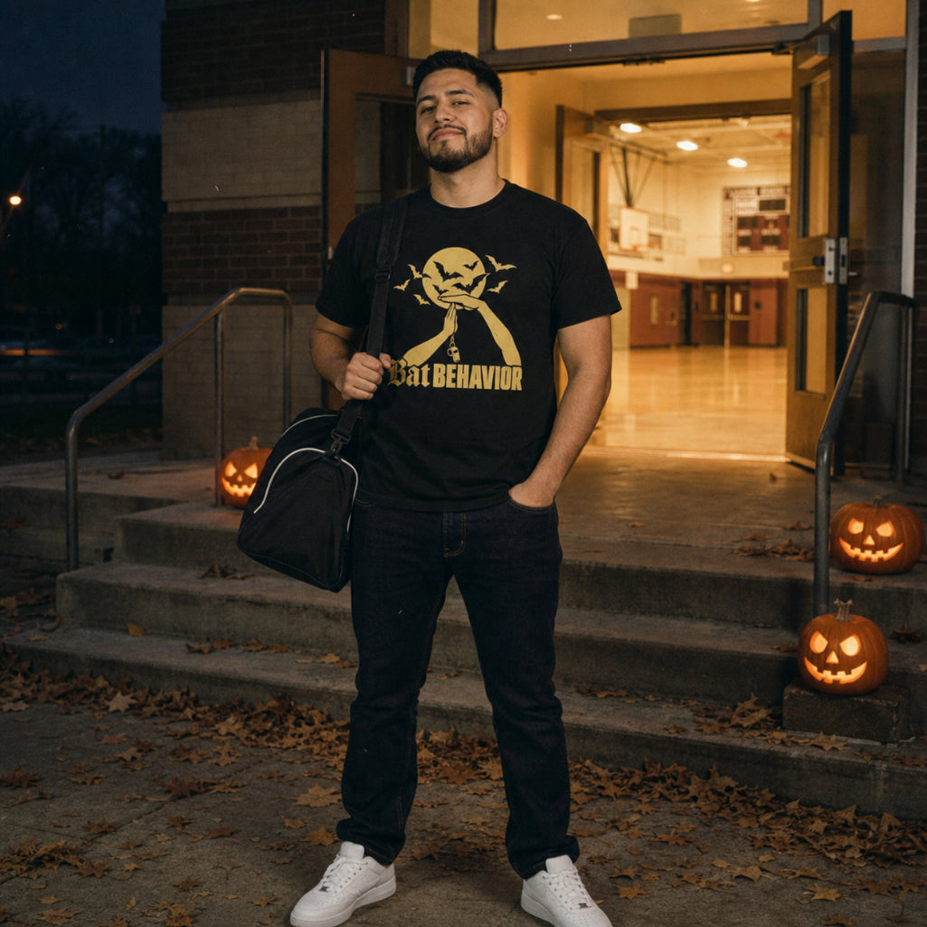 Man wearing the black Bat Behavior Tee with a gym bag outside a lit school gym entrance, jack-o'-lanterns on the steps – Ref' Crew basketball referee Halloween tee