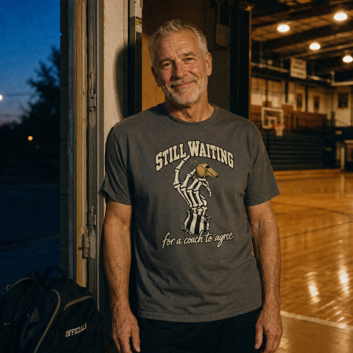 Referee wearing the Pepper Still Waiting Tee at the open door of a lit gym at dusk, officials bag at his feet – Ref' Crew basketball referee Halloween tee