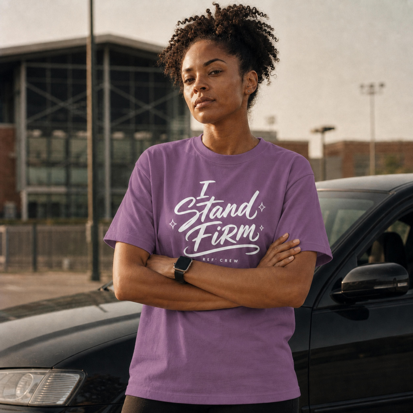 Woman wearing the Ref' Crew "I Stand Firm" tee in Berry, arms crossed outside the arena at dusk