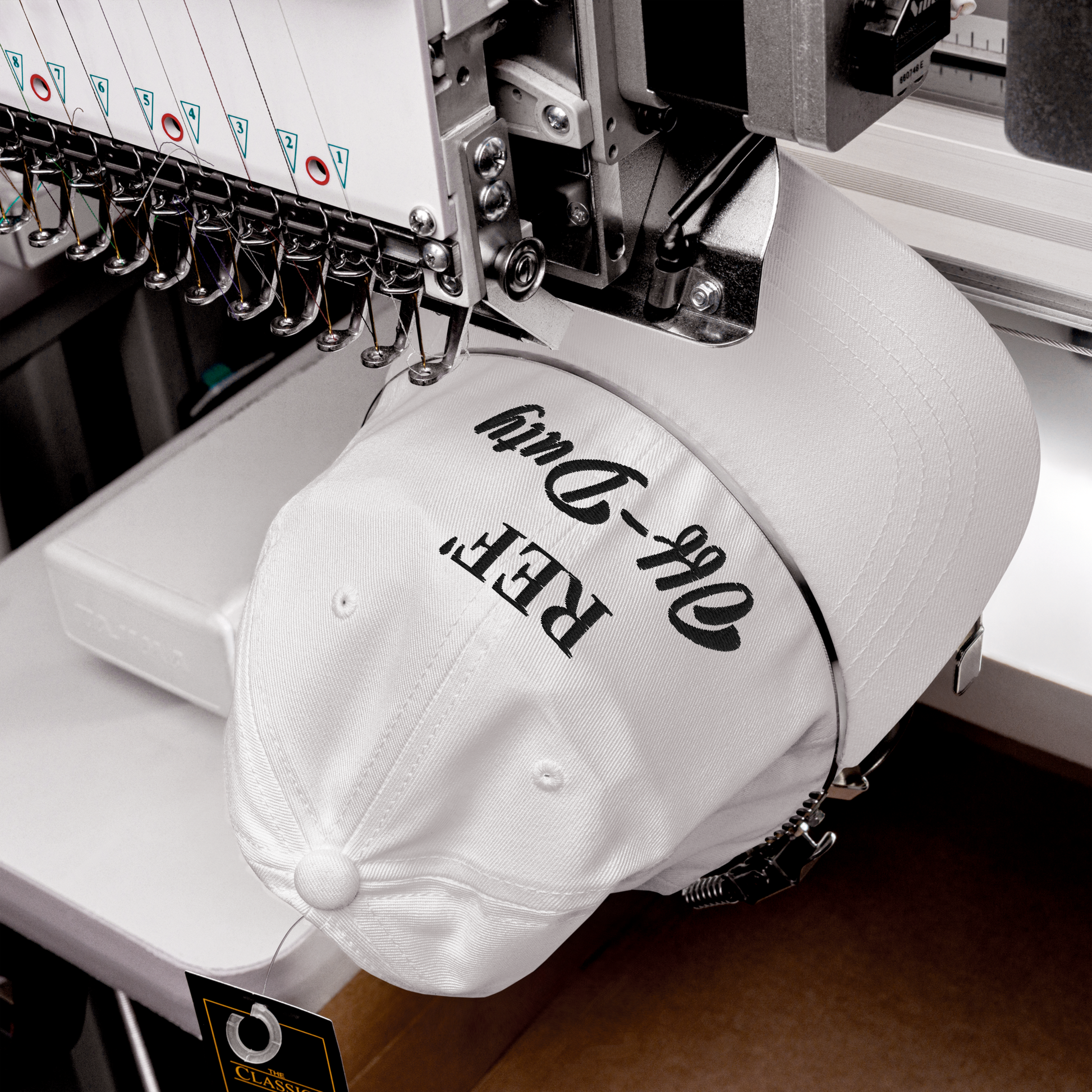 White "REF' Off-Duty" dad hat with black embroidery, being stitched on an industrial machine.